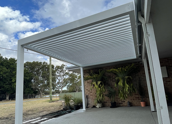 White louvre roof pergola over patio beside brick wall with hanging plants and garden view